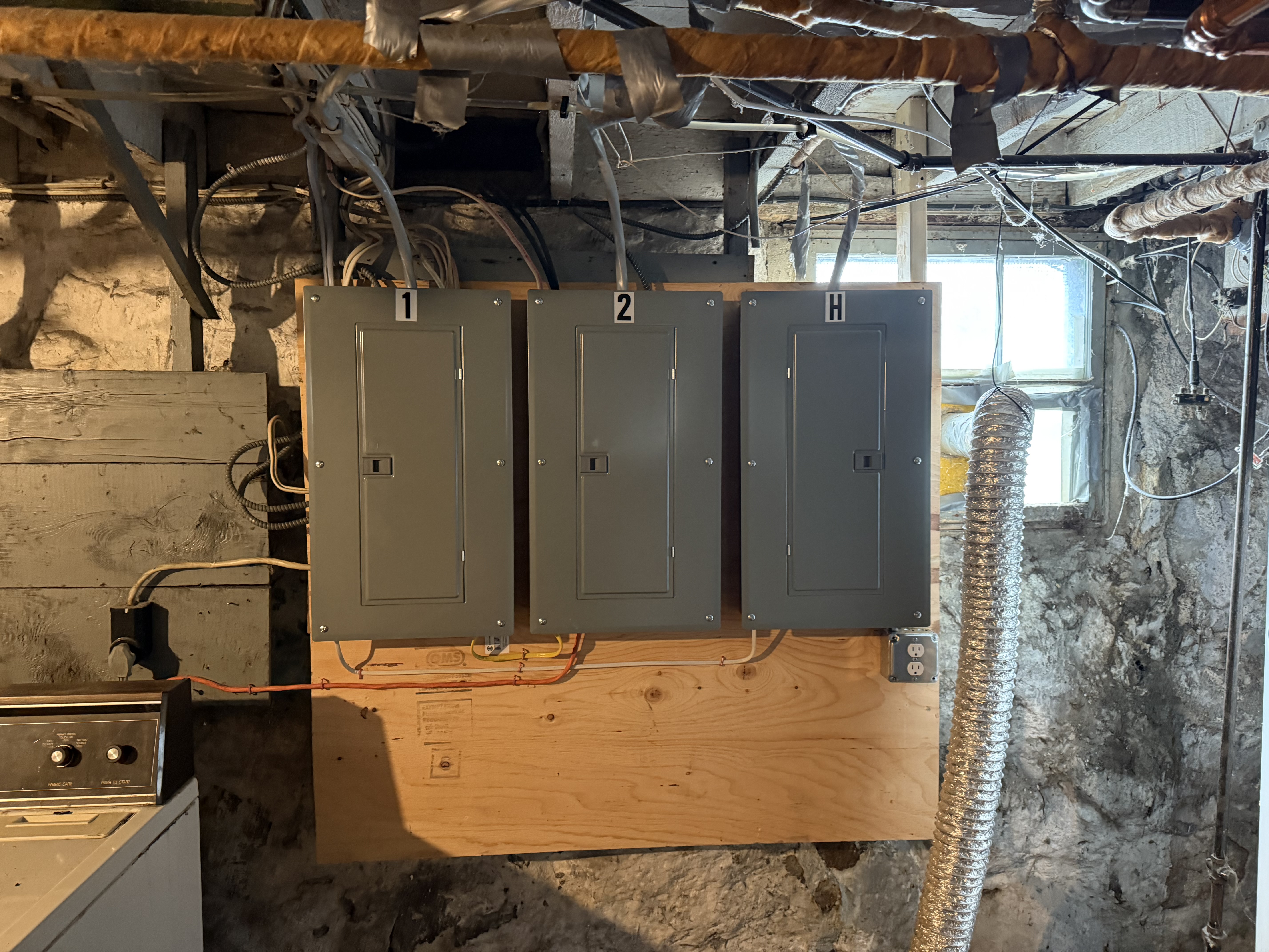 Three gray load centers labeled 1, 2 and H side by side on a plywood backboard in a stone-walled basement, dryer vent beside them, North Attleborough, MA
