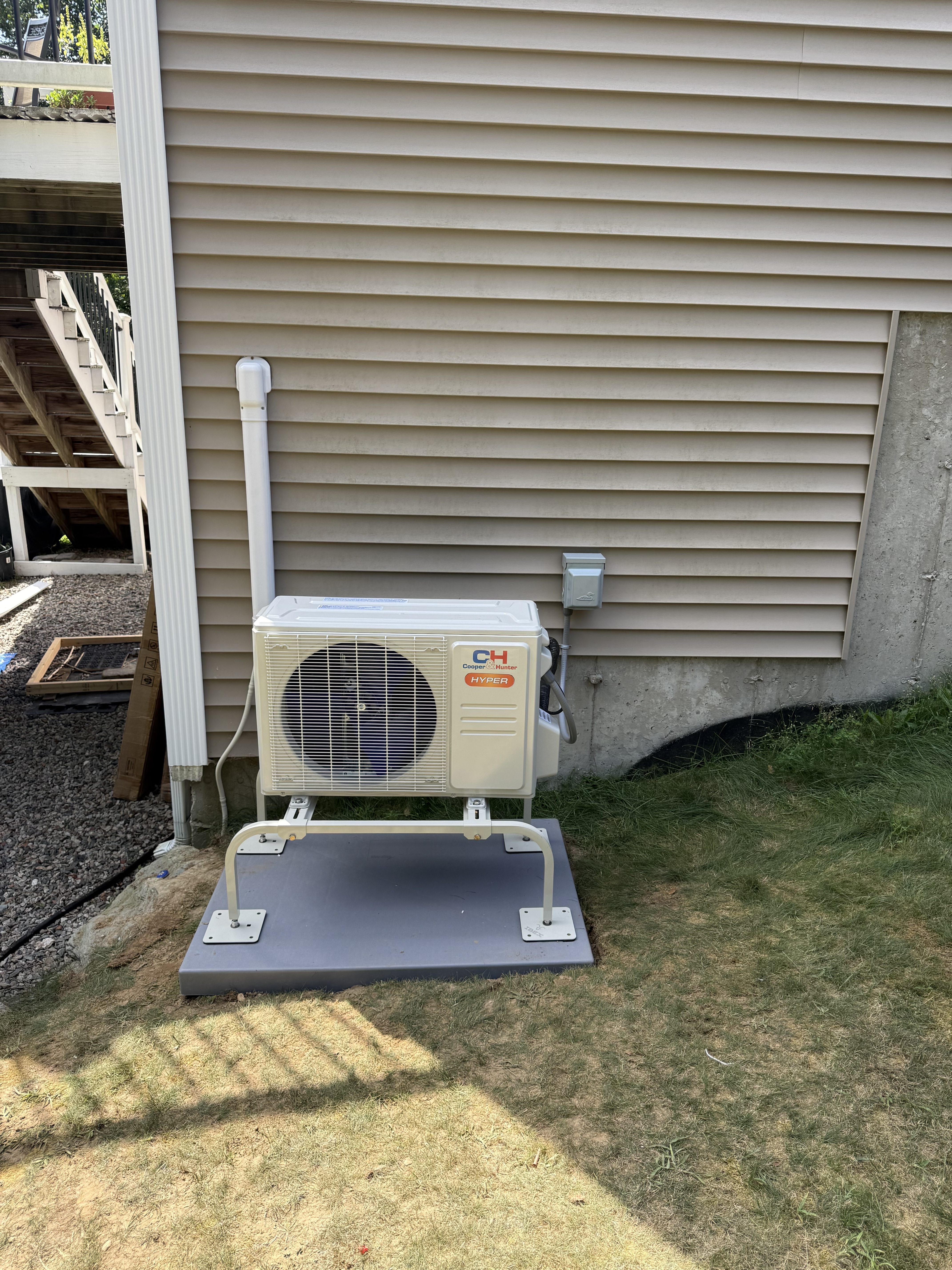Cooper and Hunter HYPER mini split condenser on a white wall stand set on a gray composite pad beside beige vinyl siding, outdoor disconnect box above it on the wall, deck stairs at left, lawn in summer sun, the kind of work we do in Raynham, MA